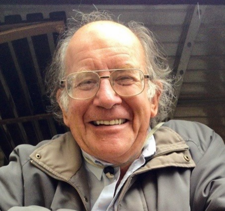 A cheery man with glasses and a brown coat smiling at the camera 